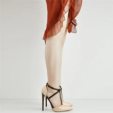 Load image into Gallery viewer, Beige high-heeled shoes with black straps worn with a rust-colored top and white pants on a light gray background.
