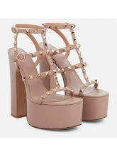 Load image into Gallery viewer, A pair of platform high-heeled sandals in a light nude pink faux leather, decorated with gold-tone metal studs and straps, shown against a plain light gray background.