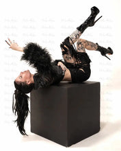 Load image into Gallery viewer, Person in black outfit with feathered elements lying on a black cube against a white background