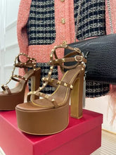 Load image into Gallery viewer, A pair of platform high-heeled sandals with chunky block heels and multiple straps adorned with gold-tone studs and buckles, displayed on a pink shoebox with a pink and black tweed jacket and a black crocodile-embossed handbag in the background.