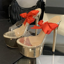 Load image into Gallery viewer, Gold high-heeled shoes with red bows on a reflective surface.