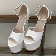 Load image into Gallery viewer, A pair of glossy white platform high heels with ankle straps and small gold-tone buckle details, displayed on a glass shelf against a neutral indoor background.