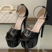 Load image into Gallery viewer, A pair of glossy black patent leather high-heeled sandals with platform soles, cutout toes, and studded heel accents, displayed on a reflective surface with a white quilted handbag in the background.