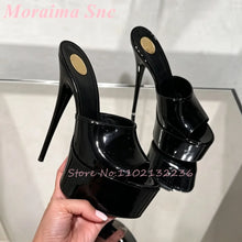 Load image into Gallery viewer, A pair of black patent leather high-heeled mule sandals with platform soles and stiletto heels, held in a hand against a light-colored tiled background.