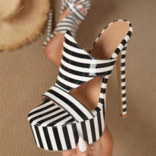 Load image into Gallery viewer, A close-up of a black and white striped high-heeled mule sandal being held by a hand, with a matching pair worn on feet in the background on a beige floor.