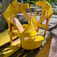 Load image into Gallery viewer, A pair of bright yellow high-heeled platform sandals with cutout mesh panels and ankle straps, photographed outdoors on a weathered yellow wooden bench with a tree and paved path in the background.