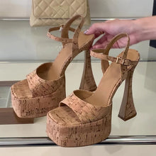 Load image into Gallery viewer, A pair of platform high-heel sandals with a cork-textured pattern in tan and brown, shown on a glass display shelf with a hand holding one shoe. A quilted beige handbag is visible in the background.
