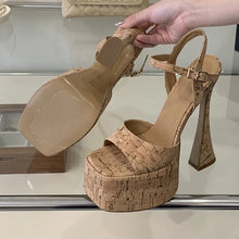 Load image into Gallery viewer, A pair of chunky platform high-heel sandals made from cork, shown with one shoe being held by a hand to display the sole and heel detail.