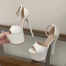 Load image into Gallery viewer, A pair of glossy white platform high-heel sandals with ankle straps and spiked heels, shown with one shoe on a glass surface and the other being held by a hand.