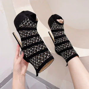Black high-heeled shoes with rhinestone embellishments held by a hand against a neutral background