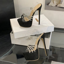 Load image into Gallery viewer, Black high-heeled shoes with clear straps on a white platform.