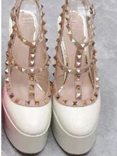 Load image into Gallery viewer, A pair of platform high-heeled shoes with glossy white patent leather toes, clear straps, and gold pyramid studs, shown from a top-down view on a gray textured surface.