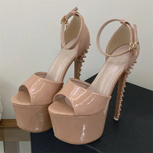 Load image into Gallery viewer, A pair of patent leather platform high heels in a nude pink color, featuring studded heels and ankle straps with gold-tone hardware, displayed on a dark surface against a plain wall.