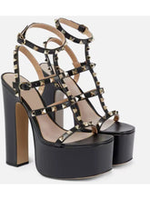 Load image into Gallery viewer, Black platform high-heeled sandals with multiple strappy ankle-strap designs and gold-tone pyramid studs, shown on a light gray background.