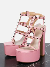 Load image into Gallery viewer, Pink platform high-heeled sandals with multiple cutout straps and pyramid stud embellishments, shown on a dark marble surface against a light gray background.