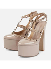 Load image into Gallery viewer, A pair of platform high-heel sandals in a glossy nude-beige patent leather, decorated with gold-toned pyramid studs along the straps and around the toe box, set against a plain light gray background.