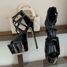 Load image into Gallery viewer, A pair of glossy black patent leather high-heeled platform sandals with crisscross ankle straps, displayed on a glass shelf next to a white quilted handbag.