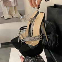 Load image into Gallery viewer, Black high-heeled sandals with clear straps held by a hand in a store setting.