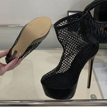 Load image into Gallery viewer, Black fishnet high-heeled shoe with a beige sole on a reflective surface.